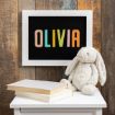 A chalkboard with the name "OLIVIA" in colorful letters is framed in white and hangs on a wooden wall. Below, a plush bunny and two stacked books sit on a small white table.