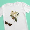 A white t-shirt with a large botanical illustration of a white magnolia flower and green leaves on the front, placed next to a pair of dark sunglasses on a light turquoise background.