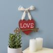 A red heart-shaped sign with the word "LOVE" and a decorative key hangs on a lace ribbon above two white candles and a green potted succulent against a blue wall.