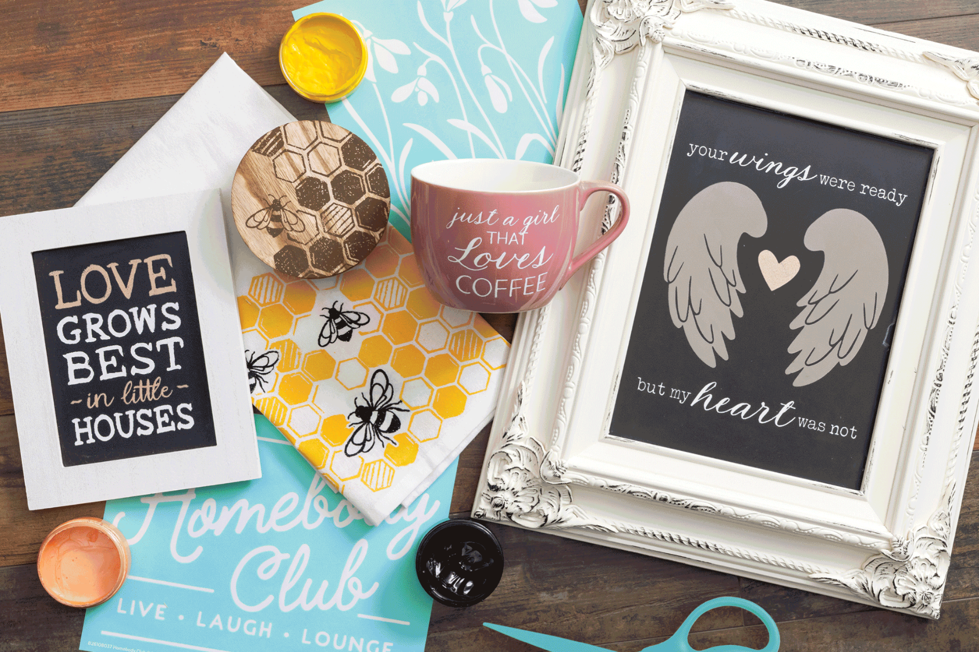 A collection of decorative items, including a framed quote with angel wings, a mug, a beehive-patterned cloth, yellow paint, scissors, and prints with inspirational sayings, arranged on a wooden surface.