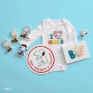 A baby onesie with “I ♥ HUGS” and Snoopy, Peanuts character cutouts, a round Snoopy sign reading “Raw Strength & Courage,” and a card with “BE” in colorful letters, all arranged on a blue background.