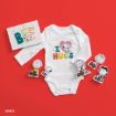 A white baby onesie with "I love hugs" and Snoopy printed on it lies on a red background, surrounded by Peanuts character cutouts and a greeting card with "BE" and colorful designs.