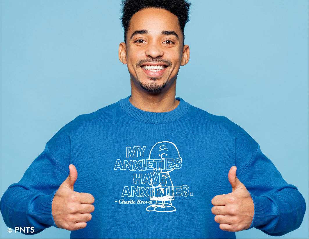 A smiling man in a blue sweatshirt gives two thumbs up. The sweatshirt features an outline drawing of Charlie Brown and the text "My anxieties have anxieties. – Charlie Brown." The background is light blue.