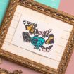 A gold ornate picture frame displays an illustration of six colorful butterflies on white paper, with shades of orange, black, blue, yellow, and pink. The frame rests on a pink and teal background.