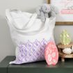 A white tote bag with purple bunny and floral patterns sits on a green table. A gray stuffed bunny peeks over the bag. Decorative egg-shaped cutouts and a painted wooden egg are nearby.