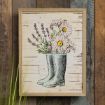 A framed illustration of green rain boots filled with daisies, lavender, and other flowers, hanging on a rustic wooden wall. Some green grass is visible in the foreground.