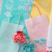 Pastel-colored Easter gift bags with bunny and egg designs, two jars of red and blue jelly beans, and decorative paper with Easter-themed text and illustrations arranged on a light surface.