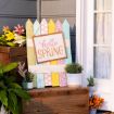 A colorful wooden sign reading “hello SPRING” is displayed outdoors among potted plants and flowers, next to a window with lace curtains and wooden crates.