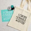 A cream tote bag with the text "1 Cross + 3 Nails = 4 Given" sits beside a turquoise stencil of the same text, a jar of black ink, and a rubber stamp on a light marble surface.