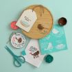 A flat lay of bird-themed crafts, including painted wooden plaques, a tea towel, stencils, scissors, twine, and containers of pink and teal paint on a light blue background.
