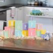Colorful pastel blocks spelling "Hoppy Easter" with bunny and egg illustrations are arranged on a wooden table with Easter grass, eggs, and a plush bunny, in a modern kitchen setting.