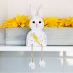 A white wooden bunny decoration with dangling legs sits on a shelf, holding a carrot. Behind it is a wooden box filled with bright yellow artificial flowers.