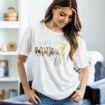 A woman with long, light brown hair smiles and looks down while wearing a white t-shirt featuring colorful birds and yellow irises. She stands in a cozy, well-lit living room with shelves and decor in the background.