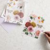 A hand places a puzzle piece onto a partially completed floral jigsaw puzzle featuring colorful flowers and a butterfly, with the puzzle box and guide sheet nearby on a light, textured surface.