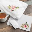 A white blanket and a white mouse pad, both with pink floral designs, are on a wooden surface next to a white cup filled with tea and a white wireless computer mouse.