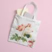 A white tote bag with pink and white roses printed on it lies on a pink surface. Two books, one cream and one gray, are partially tucked inside the bag.