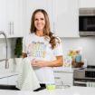 A woman smiles while drying a dish with a striped towel in a modern kitchen. She wears a white shirt that says "Lilly Rabbit Easter is for Jesus." There are Easter decorations and a bowl on the counter.