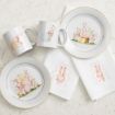 Easter-themed tableware set featuring plates, mugs, and napkins decorated with pastel illustrations of bunnies holding colored eggs, placed on a light marble surface.