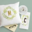 A white pillow with a gold letter "M" and leafy design, a white napkin with a gold letter "C" and leaves, a cup of tea, an open book, and eyeglasses on a pale green surface.