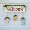 A decorative sign with the word "Welcome" hangs on a wall, adorned with green moss and three small, colorful birdhouses below, each also topped with moss. The background is light blue tiles.