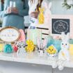 A countertop decorated with Easter-themed items, including paper cutouts of bunnies, sheep, and a chick, colorful eggs, jars of candy, pastel decorations, and framed art. Two people stand in the background holding drinks.
