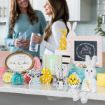 Two women stand and chat in a kitchen decorated for Easter with pastel-colored bunny, sheep, chick, and egg crafts, jars of candy, and framed art with spring themes arranged on the counter.