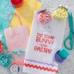 Colorful Easter-themed items on a countertop, including a towel with "Did Some Bunny Say Easter" and bunny graphics, pastel gift bags with bunnies, jars of candy, scissors, and purple crafting paint.
