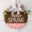 A round wooden sign with “hello SPRING” painted in white, decorated with pink polka dots, white flowers, and a large white and striped bow, hanging on a white door.