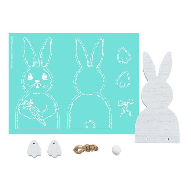 A DIY bunny craft kit including a turquoise stencil sheet with bunny and accessory outlines, a white wooden bunny cutout, two wooden feet, a small rope, a white pompom, and a bow shape.