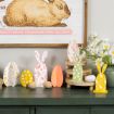 A green table displays colorful wooden bunny and egg decorations, wooden eggs, and a vase of daisies. Above is wall art featuring a brown rabbit and candy names on a pink banner.