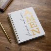A spiral-bound notebook on a wooden table with gold text reading "AMEN" and black cursive text reading "forever and ever" on the cover. A gold pen and part of a book are nearby.