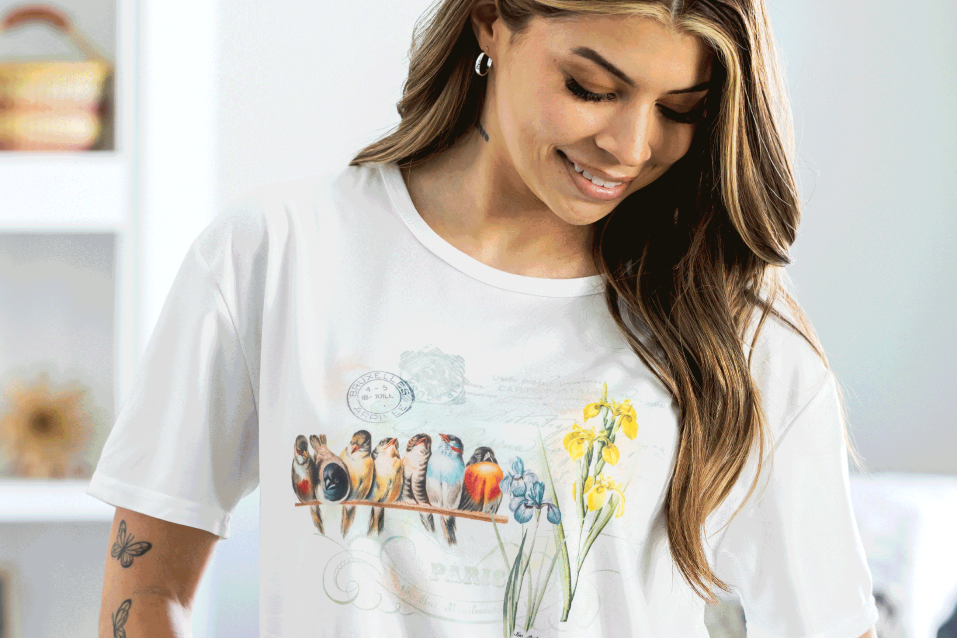 A woman with long hair smiles while looking down at her white t-shirt, which features an illustration of birds on a branch and yellow flowers. She has a butterfly tattoo on her arm and is indoors.