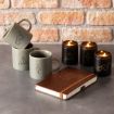 Three gray mugs with Harry Potter-themed designs, three black candles with similar designs, and a brown notebook are arranged on a gray surface in front of a brick wall.
