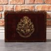 A vintage-style suitcase with brass accents and a gold Hogwarts crest, featuring the letter "H" and the motto "Draco Dormiens Nunquam Titillandus," sits on a grey surface in front of a brick wall.