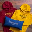 Two hoodies, one red with "Gryffindor Seeker" and one yellow with "Hufflepuff Prefect," are laid on a wooden floor next to a blue pillow reading "Ravenclaw Alumni.