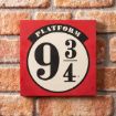 A square sign with a red border and a white circle in the center reads “PLATFORM 9 3/4” in black text, mounted on a brick wall.