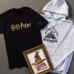 A black Harry Potter T-shirt, a gray hoodie with “Waiting for my letter to Hogwarts” text, and a framed illustration of the Sorting Hat on a stack of books are arranged on a gray surface.