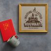 A framed print reads "Waiting on my letter to Hogwarts Owl Post Service" with a castle, owl, and envelope. Next to it are a red notebook with a lion emblem and a gray mug with a triangular symbol, all on a gray surface.
