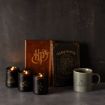 A set of black candles with gold Harry Potter symbols, two Hogwarts-themed notebooks, and a gray mug with the Deathly Hallows symbol are arranged on a gray surface against a dark background.
