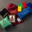 A flat lay of Harry Potter-themed merchandise, including T-shirts, hoodies, pillows, notebooks, a framed print of the sorting hat, and a keychain, each representing Hogwarts houses.