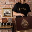 A person wearing a black Harry Potter shirt holds a large brown Hogwarts suitcase. On a table are themed candles, notebooks, and mugs. A framed Hogwarts print hangs on a brick wall in the background.