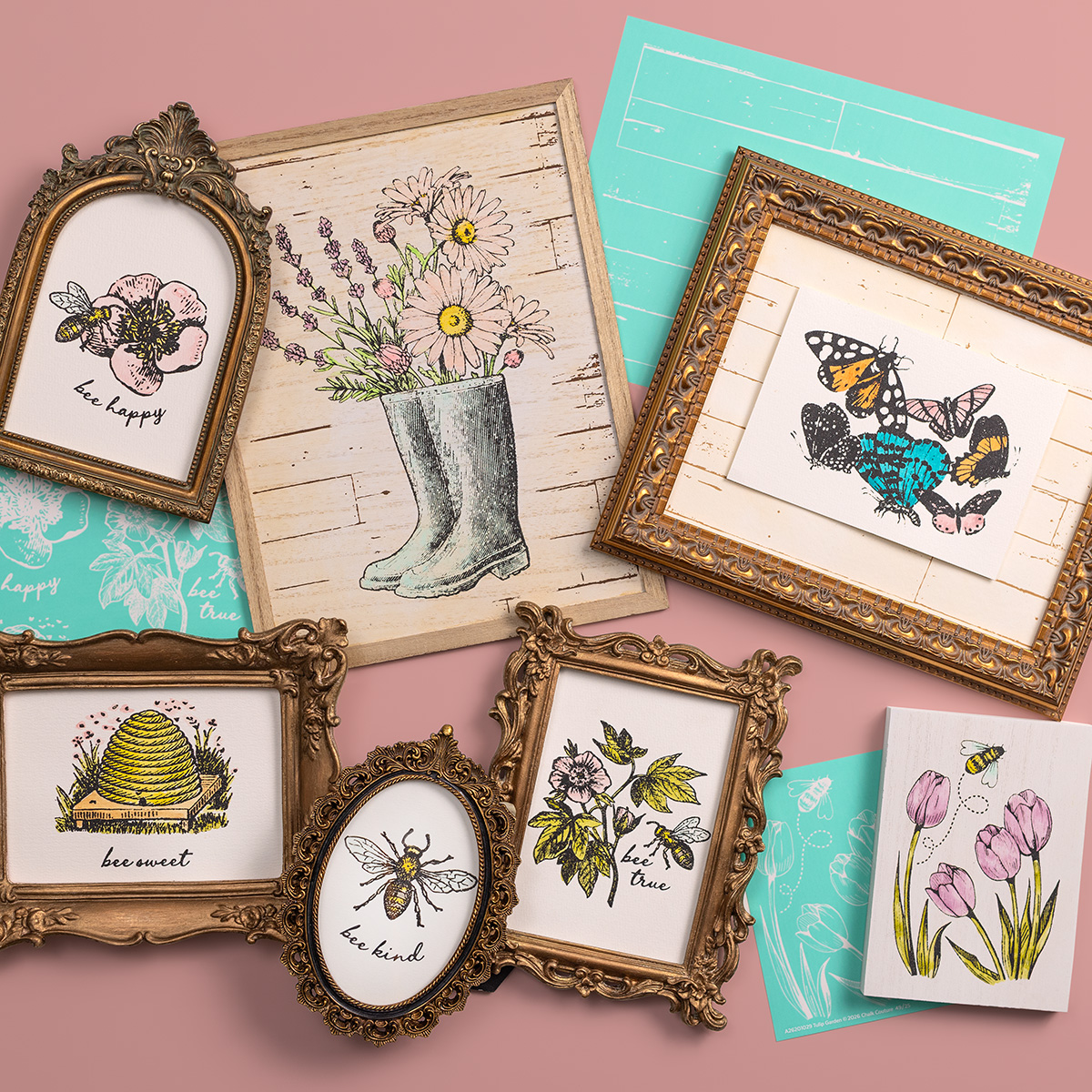 A flat lay of framed artwork on a pink surface, featuring floral designs, bees, butterflies, rubber boots with flowers, and handwritten words like "be happy," "be sweet," "be kind," and "bee true." Blue stencils are also visible.