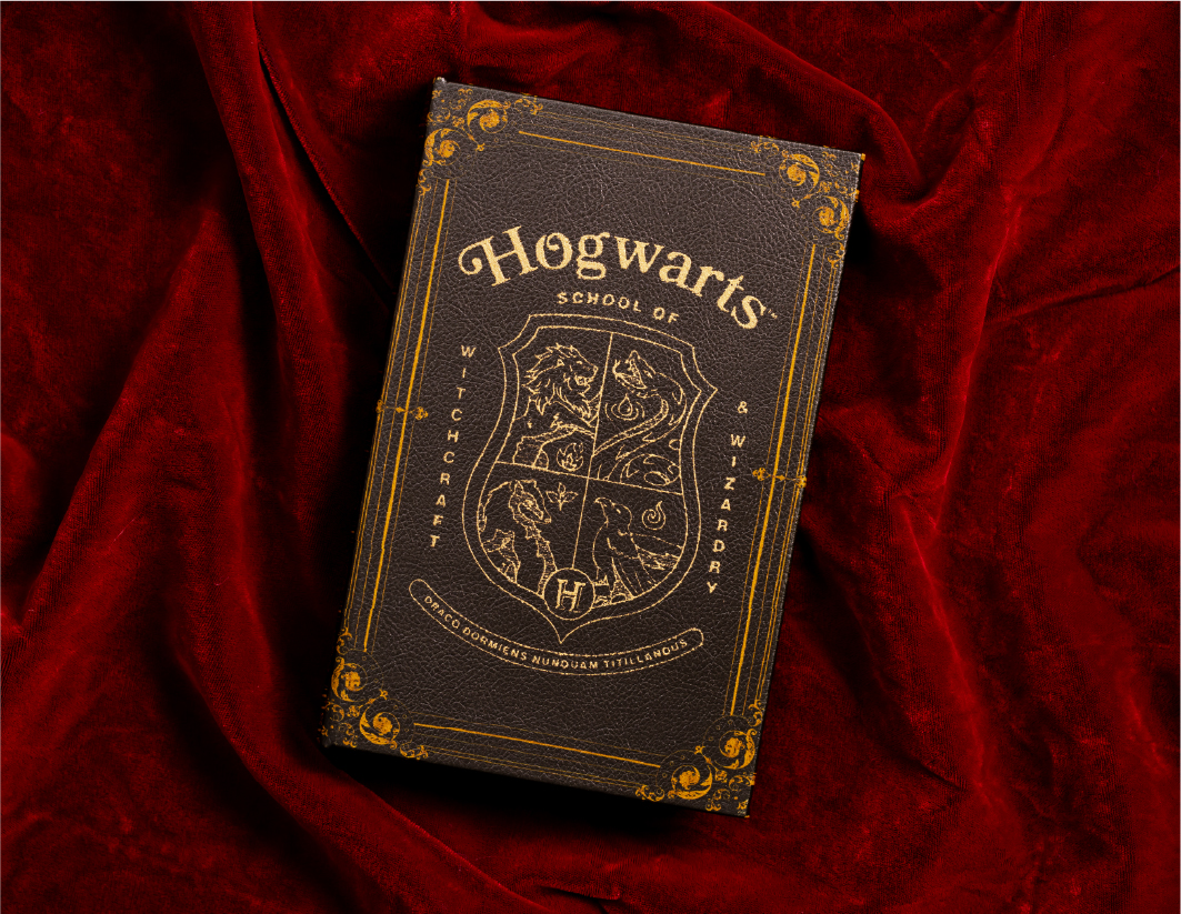 A hardcover book titled "Hogwarts School of Witchcraft & Wizardry" with a gold-embossed crest rests on red velvet fabric. The cover features ornate borders and house symbols from the Harry Potter series.