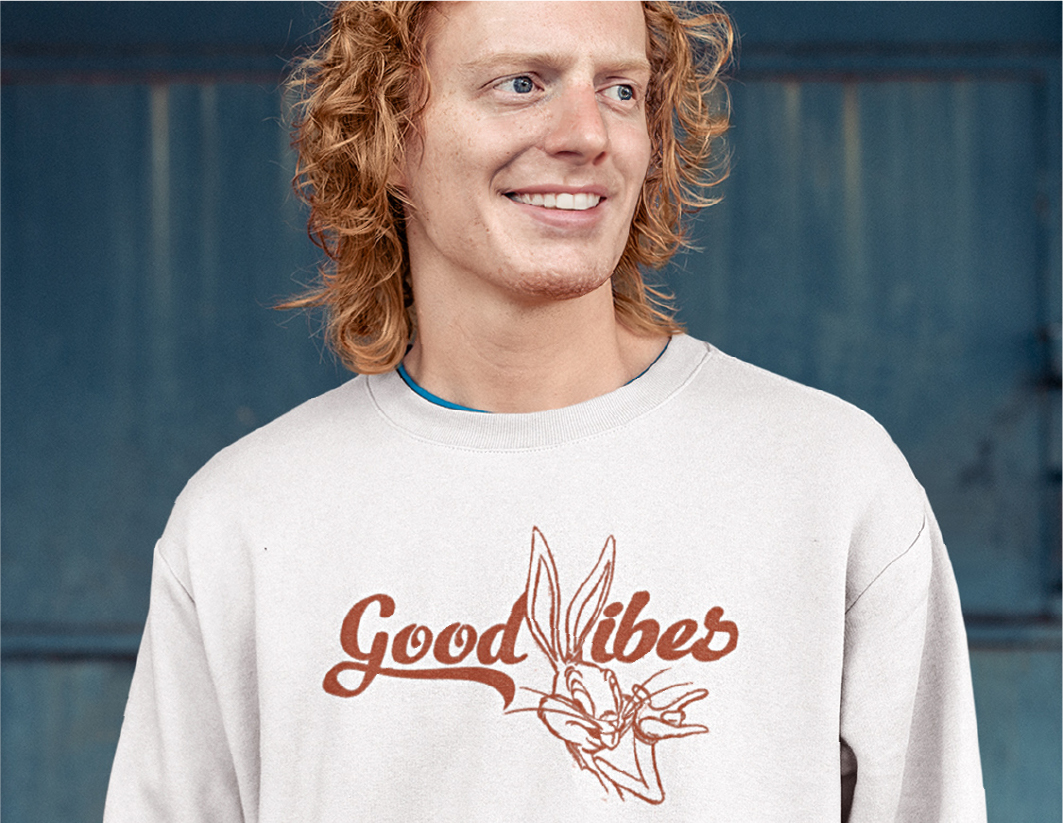 A smiling person with curly hair wears a light-colored sweatshirt featuring the words “Good Vibes” and an illustration of Bugs Bunny making a peace sign. The background is a blue, textured wall.
