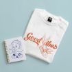 A white t-shirt with an orange “Good Vibes” text and Bugs Bunny graphic lies next to a spiral notebook with a blue line drawing of Donald Duck holding a mug and plate, all on a light blue background.