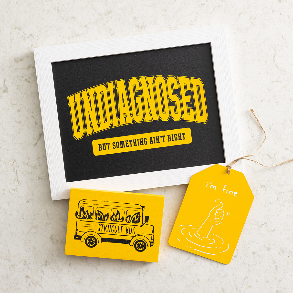 A framed sign reads "Undiagnosed but something ain't right." Below it are a yellow magnet with a burning bus labeled "Struggle Bus" and a yellow tag showing a hand giving a thumbs-up from water, captioned "i'm fine.