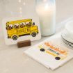 A card and napkin both display a yellow school bus with flames in its windows and the words "STRUGGLE BUS." The items are placed on a countertop next to a lit candle, stacked plates, and a bowl.