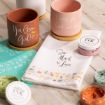 Three pastel plant pots with positive messages, ink containers, a stencil, and a white tea towel with floral designs and "Sow Much Love" text are arranged on a light wooden surface.