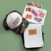 A trucker hat reading "TIRED 24/7," a white pouch decorated with colorful stickers and filled with markers, and a small bag labeled "WOW! WOW!" are arranged on a green background.