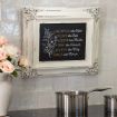 A framed chalkboard on a tiled kitchen wall lists virtues with names: "Sacrifice like Esther, Love like Ruth, Serve like Martha, Pray like Hannah, Believe like Mary, Dance like Miriam." A pot and vase of flowers are nearby.